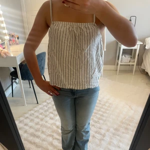 Randig linnetopp från AWARE, S - Säljer en luftig och snygg linnetopp från AWARE i storlek S. Toppen är vit med svarta vertikala ränder, har smala axelband och rak passform. Perfekt för varma dagar och enkel att matcha med jeans eller kjol.