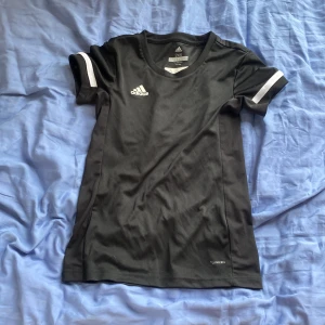 Svart Adidas fotbollströja 2XS - Snygg svart fotbollströja från Adidas i storlek 2XS. Tröjan har vita detaljer på ärmarna och är tillverkad i lätt och ventilerande ClimaCool-material som passar perfekt för träning. Klassisk Adidas-logga på bröstet och sportig passform.