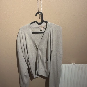 Grå/beige  kofta med fickor och knapp - Snygg grå kofta med v-ringning och en stor knapp framtill. Modellen är lite loose och har två praktiska fickor framtill. Perfekt att slänga över en t-shirt eller linne. Mjuk och skön, passar till många olika outfits.