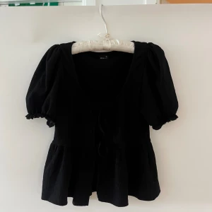 Svart blus med puffärm och knyt - Supersöt svart blus med puffiga korta ärmar, djup v-ringning och knyt framtill. Blusen har en volangkant nedtill och är tillverkad i ett lätt, strukturerat tyg som ger en luftig känsla. Perfekt för dig som gillar romantiska detaljer och trendig siluett. Skicka ett meddelande för fler bilder.