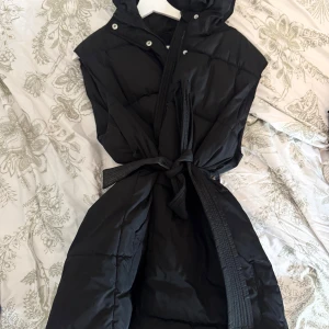 Svart quiltad dunväst med luva - Svart dunväst med quiltad design och huva. Västen har knytband i midjan för en snygg siluett och stängs med både dragkedja och tryckknappar. Perfekt för lager-på-lager