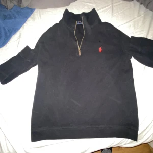 Svart half zip tröja från Polo Ralph Lauren - Svart långärmad tröja från Polo Ralph Lauren med half zip och klassisk röd logga broderad på bröstet. Tröjan har hög krage och är tillverkad i mjuk bomull, perfekt för lager-på-lager. Enkel och stilren design som funkar till det mesta.