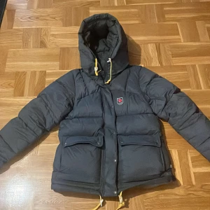 Grå dam Fjällräven Expedition dunjacka S - Grå dunjacka från Fjällräven Expedition Series 1974, dammodell i storlek S. Jackan har stor huva, gula dragskor, två stora fickor framtill och klassisk Fjällräven-logga på bröstet. Perfekt för kalla dagar med sin fluffiga och quiltade design.