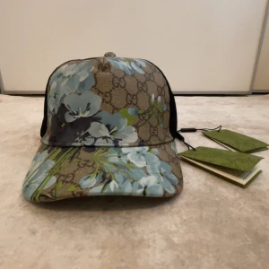 Gucci keps med blå blommor - Snygg keps från Gucci med klassiskt GG-mönster i beige och brunt, dekorerad med stora blå och ljusgröna blommor. Baksidan är i svart mesh för extra ventilation. Perfekt statement-piece för dig som vill sticka ut.