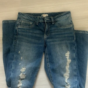 Blå bootcut jeans från Gina Tricot - Snygga blå jeans från Gina Tricot med bootcut-modell och slitna detaljer framtill. Klassisk femficksdesign, markerade sömmar och coola bakfickor med broderi. Perfekta för dig som gillar en avslappnad och trendig look.
