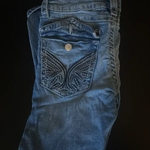 Blå jeans med broderad bakficka - Snygga blå jeans med cool broderad detalj på bakfickan och klassisk femficksdesign. Jeansen har en normal passform och är tillverkade i mjukt denimtyg. Perfekta för dig som gillar unika detaljer och en avslappnad stil.