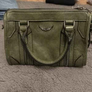 Zadig voiltare olivgrön - Cool olivgrön axelväska i skinnimitation med silverfärgade detaljer och ett emblem med glittrande vingar framtill. Väskan har både handtag och lång justerbar axelrem samt dragkedja upptill och dekorativa dragkedjor på framsidan. (Köptes på plick)