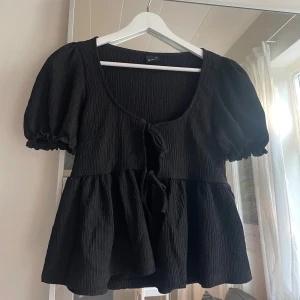 Svart blus med knyt från Gina Tricot - Supersöt svart blus från Gina Tricot i storlek XS. Modellen har puffiga korta ärmar, knyt framtill och en flowy, volangig nederdel. Tyget är strukturerat och ger en trendig look. Perfekt för dig som gillar en feminin och chill stil.