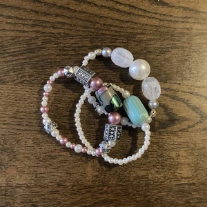 Tre pärlarmband med färgglada detaljer - Säljer tre unika armband med pärlor i vitt, rosa och silver. Ett armband har stora vita och genomskinliga pärlor, ett har en turkos pärla med guldiga ränder och ett har rosa pärlor, silvriga detaljer och en cool glaspärla med svart och grön effekt. Perfekt för dig som gillar att mixa och matcha smycken.