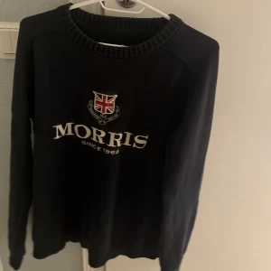 Svart stickad tröja Morris S - Svart stickad tröja från Morris med broderad logga och brittisk flagga på bröstet samt siffran 68 på ena ärmen. Klassisk rund halsringning och ribbade muddar. Perfekt för dig som gillar preppy stil.