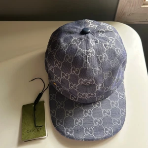 Blå mönstrad keps från Gucci - Snygg blå keps från Gucci med klassiskt GG-mönster över hela kepsen. Kepsen har böjd skärm och är tillverkad i bomullsmaterial. Märkets logga syns både på utsidan och insidan. Perfekt för dig som vill ha en lyxig och trendig accessoar.