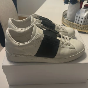 Valentino Garavani Open sneakers vit/mörkblå - Snygga Valentino Garavani Open sneakers i vitt skinn med bred svart läderdetalj över snörningen. Klassisk rund tå, vita skosnören och diskreta nitar på sulan. Kommer med originalkartong och dustbag. Perfekta för dig som gillar lyxiga och stilrena sneakers.