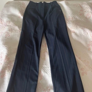 Mörkblå randiga kostymbyxor Nümph - Snygga mörkblå kostymbyxor från Nümph med tunna vertikala ränder. Byxorna har hög midja och raka ben med klassisk passform. Perfekta för en stilren look och lätta att matcha med olika toppar.