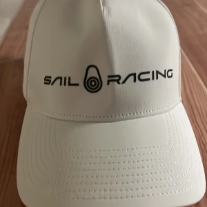 Vit keps från Sail Racing - Vit keps från Sail Racing med svart logotyp framtill. Kepsen har böjd skärm, justerbart spänne i metall baktill och är tillverkad i polyester och elastan för en sportig känsla. Perfekt accessoar för en clean och stilren look.