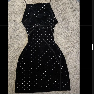 Svart sammetsklänning med prickar - Supersnygg svart sammetsklänning med prickiga detaljer och tunna axelband. Klänningen har en tight passform och är kort, med en öppen rygg i mesh som ger en extra cool detalj. Perfekt för dig som vill sticka ut med en klassisk men trendig look.