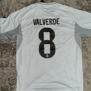 Real Madrid Valverde 8 Adidas tröja L - Säljer en officiell Real Madrid fotbollströja från Adidas i storlek L. Tröjan är vit med svarta detaljer, gula linjer och grå partier på sidorna. På ryggen står Valverde och nummer 8. Framsidan har klubbmärke och Emirates Fly Better-tryck. Materialet är lätt och andas.
