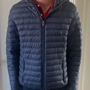 Blå dunjacka från JOTT med huva - Snygg blå dunjacka från JOTT med huva och horisontella sömmar. Jackan har dragkedja framtill och en patch med JOTT-logga på ärmen. Perfekt för kyliga dagar och enkel att matcha med olika outfits.