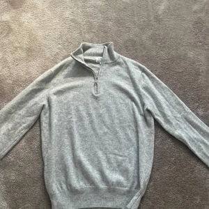 Grå halvzip stickad tröja från S&T studios - Snygg ljusgrå stickad tröja från S&T Studios med halv dragkedja i halsen. Tröjan har långärmade ribbade muddar och en klassisk, clean look som funkar till jeans eller chinos. Perfekt för dig som gillar stilrena och bekväma plagg.