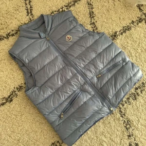 Ljusblå dunväst från Moncler - Snygg ljusblå dunväst från Moncler med klassisk logga på bröstet. Västen har två dragkedjeförsedda fickor framtill och stängs med dragkedja. Perfekt för lager-på-lager och ger en clean look till din outfit.