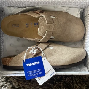 Beige Birkenstock Boston i mocka - Säljer ett par snygga Birkenstock Boston i beige mocka med klassisk korksula och justerbart spänne i bronsfärg. Skorna har en rund tå och platt sula, perfekt för chill vardagsstil. Ikonisk Birkenstock-logga på sidan och mjuk innersula för extra komfort.