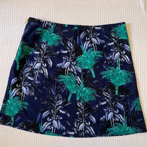 Mönstrad kjol Holly & Whyte Lindex - Snygg kortkjol från Holly & Whyte by Lindex i marinblått med grönt och vitt palmbladsmönster. Kjolen har en guldig dragkedja i sidan och är tillverkad i ett stadigt bomullstyg. Perfekt för dig som vill sticka ut med tropiska vibes.