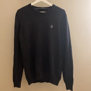 Svart stickad tröja från Sail Racing - Svart stickad tröja från Sail Racing med rund halsringning och diskret logga på bröstet. Tröjan har ribbade muddar vid ärmslut och nederkant, samt långärmad passform. Perfekt för dig som gillar stilren och enkel design.