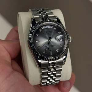 Seikmod Day-Date silverklocka diamond dial arabic - Stilren Seikmod Day-Date klocka med grå urtavla, silverfärgat armband i rostfritt stål och markerade index med stenar. Klockan har dag- och datumvisning samt räfflad bezel. Perfekt accessoar för dig som gillar klassisk och lyxig stil.