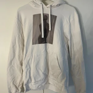 Vit hoodie med konstmotiv - Vit hoodie med tryck av konstnärlig siluett på bröstet. Tröjan har huva med snörning och en stor magficka framtill.