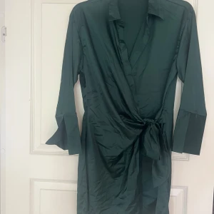 Mörkgrön omlottklänning med knyt - Snygg mörkgrön omlottklänning med långa ärmar och knyt i midjan. Klänningen har en elegant krage och är tillverkad i ett glansigt material som ger en lyxig känsla. Perfekt för dig som vill ha en stilren och trendig look.