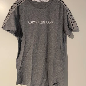 Grå t-shirt Calvin Klein Jeans XS - Snygg grå t-shirt från Calvin Klein Jeans i bomull med logga tryckt på bröstet och coola band med text längs axlarna. Klassisk rund hals och korta ärmar. Perfekt för dig som gillar stilrena och moderna plagg med en sportig touch.