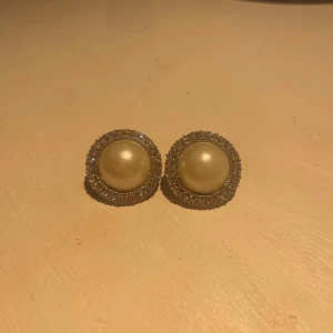Runda pärlörhängen med strasskant - Snygga örhängen med rund form, stor pärla i mitten och glittrande strass runt kanten. Färgen är vit och silver, vilket ger en elegant vibe. Perfekta om du vill addera lite glam till din look. 