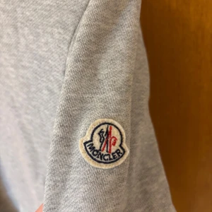 Grå maglia från Moncler  - Säljer en äkta grå kofta modell maglia från Moncler med ståkrage och hel dragkedja framtill. Koftan har två fickor med randiga dragkedjor och broderad Moncler-logga på ärmen. Ribbad mudd vid ärmslut och nederkant. Har en liten skavanka på vänster baksida, som är professionellt sydd av en skräddare finns också två effekter som du kan se på dom sista bilderna men det syns inte när du har på den! Finns här att skriva till om det är något mer du vill veta🌟 