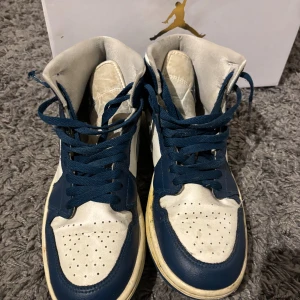 Nike Air Jordan 1 Mid blå/vit - Säljer ett par Nike Air Jordan 1 Mid i blått och vitt skinn med klassisk Swoosh och Air Jordan-logga på sidan. Skorna har perforerad tå, blå snörning och en retro vibe. Perfekta för dig som gillar streetstyle och sneakers med ikonisk design. Fint skick. Följer med box.