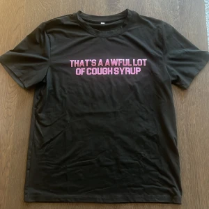 Svart t-shirt med tryck - Svart t-shirt med korta ärmar och ett stort rosa tryck på både fram- och baksidan. På framsidan står det 'THAT'S A AWFUL LOT OF COUGH SYRUP' och på baksidan finns en stor grafisk bild med samma text i rosa. 