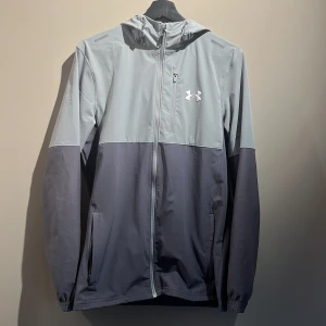 Grå vindjacka från Under Armour storlek S - Snygg tvåtonad grå vindjacka från Under Armour med huva och hel dragkedja framtill. Jackan har lång ärm, praktiska fickor och en vit logga på bröstet. Perfekt för dig som gillar sportig stil och vill ha något lätt och funktionellt. Jättebra skick( nypris 1600) säljs även inte längre.