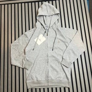 Grå hoodie från Moncler med dragkedja - Snygg ljusgrå hoodie från Moncler med huva och hel dragkedja framtill. Tröjan har Moncler-logga på bröstet, ribbade muddar och fickor på sidorna. Perfekt för chill dagar och enkel att matcha med jeans eller joggers.