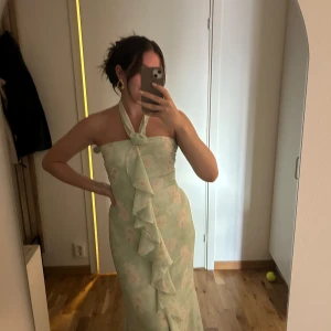 Mintgrön adoore långklänning med volang - Superfin långklänning i mintgrönt med blommigt mönster och volang framtill. Halterneck-modell som knyts i nacken och har en snygg slits. Perfekt för dig som vill sticka ut med en romantisk och trendig look.