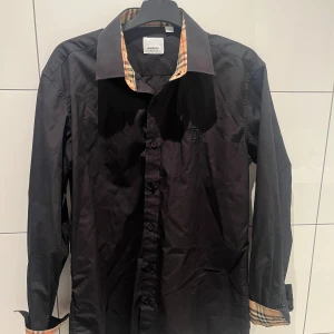 Svart Burberry skjorta med rutiga detaljer - Svart skjorta från Burberry i bomull med klassiska rutiga detaljer på insidan av kragen och ärmsluten. Skjortan har knappar framtill, lång ärm och broderad B-logga på bröstet. Snygg och stilren modell med normal passform.