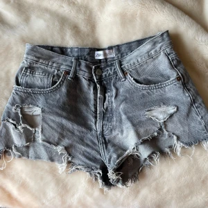 Grå jeansshorts med slitningar från Zara - Säljer ett par grå jeansshorts från Zara i storlek 36. De har hög midja, råa och fransiga kanter samt slitna detaljer med hål framtill. Klassisk femficksmodell med knapp och dragkedja. Perfekt för dig som gillar en edgy och avslappnad stil.