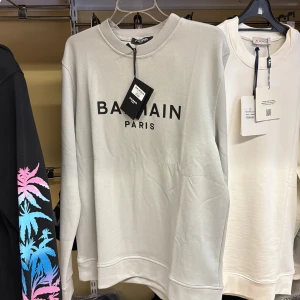 Ljusgrå sweatshirt från Balmain Paris - Snygg ljusgrå sweatshirt från Balmain Paris med svart logotyptryck på bröstet. Tröjan har rund hals och är tillverkad i 100% bomull, vilket gör den mjuk och skön. Perfekt för dig som gillar stilrena och trendiga plagg med exklusiv känsla.