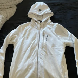 Vit hoodie Sail Racing med dragkedja - Vit hoodie från Sail Racing med hel dragkedja framtill och en extra bröstficka med dragkedja. Tröjan har huva med broderad logga och långa ärmar. 