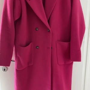 Rosa kappa från Monki i storlek S - Säljer en snygg rosa kappa från Monki i storlek S. Den har dubbelknäppning med svarta knappar, stora fickor framtill och en klassisk krage. Materialet är ullblandning som känns mjukt och varmt, perfekt för kyliga dagar.