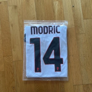 AC Milan Modric #14 vit matchtröja Puma - AC Milan fotbollströja med Modric och nummer 14 på ryggen. Tröjan är vit med svarta och röda detaljer, svart krage och logga från Puma. Materialet är lätt och andas, perfekt för match eller träning. Emirates Fly Better tryck på bröstet och klubbmärke med stjärna.