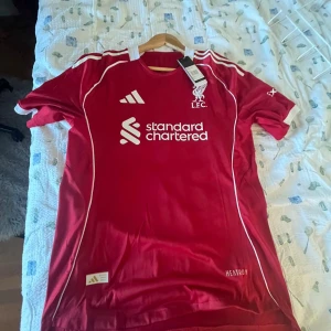Liverpool FC röd Adidas fotbollströja L - Liverpool FC officiell matchtröja från Adidas i rött med vita detaljer och logga på bröstet. Tröjan har korta ärmar, rund hals och är tillverkad i lätt, ventilerande polyester. Klassiska Adidas-ränder på axlarna och tryckt sponsor på framsidan.