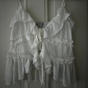 Vit volangblus från Amisu - Supersöt vit blus från Amisu med tunna axelband, volanger och knytning framtill. Plisserad struktur.