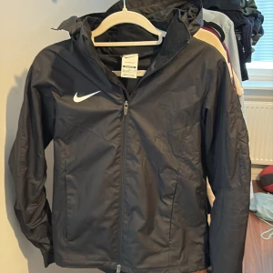 Svart Nike vindjacka Storm-FIT xs - Svart Nike vindjacka i Storm-FIT material med huva och dragkedja framtill. Loose fit-modell, passar dig som är 158-170 cm. Snygg och enkel design med vit Nike-logga på bröstet. Perfekt för blåsiga dagar och utomhusaktiviteter.