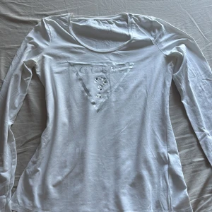 Vit långärmad tröja från Guess - Vit långärmad tröja från Guess med klassisk triangellogga framtill i silver. Tröjan har rund halsringning och är tillverkad i mjuk bomull, perfekt för en clean och stilren look.