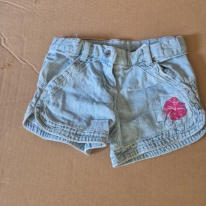 Ljusblå jeansshorts från Mexx barn - Säljer ett par ljusblå jeansshorts från Mexx i bomull. Shortsen har klassisk femficksdesign, bälteshällor och en broderad rosa blomma framtill. Perfekta för varma dagar och har en avslappnad passform.