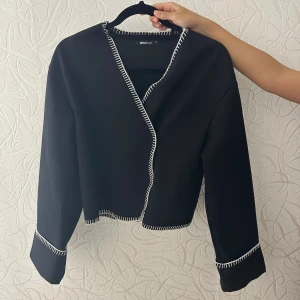 Svart kofta från Gina Tricot - Svart kofta från Gina Tricot med vit dekorativ söm längs kanterna och ärmsluten. Modellen är croppad och har lång ärm. Perfekt för lager-på-lager och enkel att styla till olika looks.