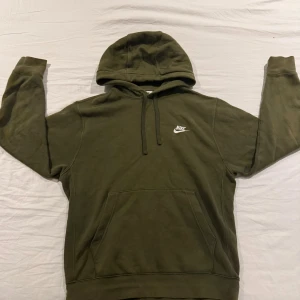 Olivgrön Nike hoodie med huva - Olivgrön hoodie från Nike storlek S (men jag som har M pasar den även)med klassisk logga på bröstet. Tröjan har huva med snörning, magficka och långa ärmar. Mjuk insida och ribbade muddar vid ärmslut och nederkant. Perfekt för chill eller streetwear.fråga gärna frågor om ni undrar nåt 